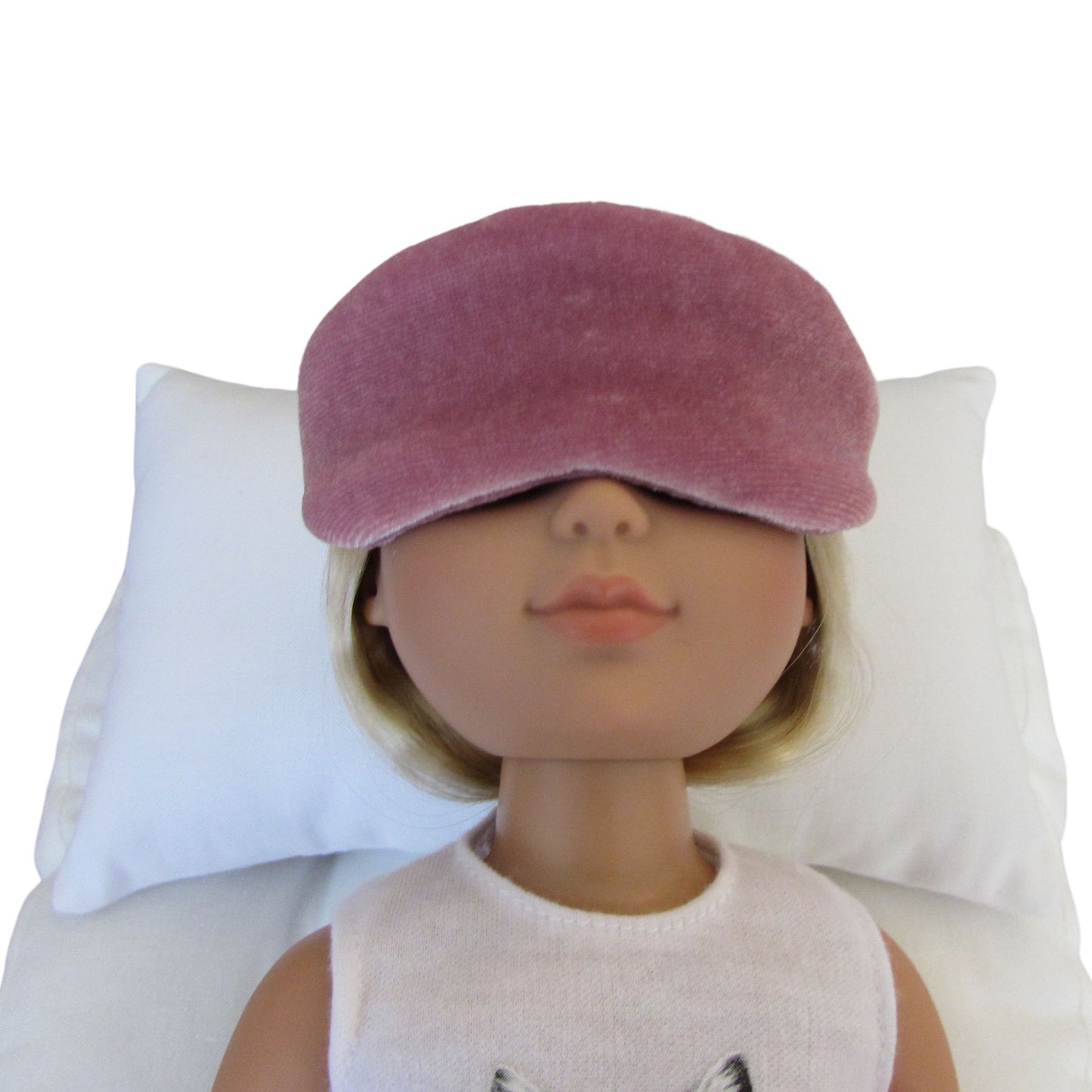 A doll with blonde hair wearing a dusty rose sleep mask. Lying on a white pillow against a white background.