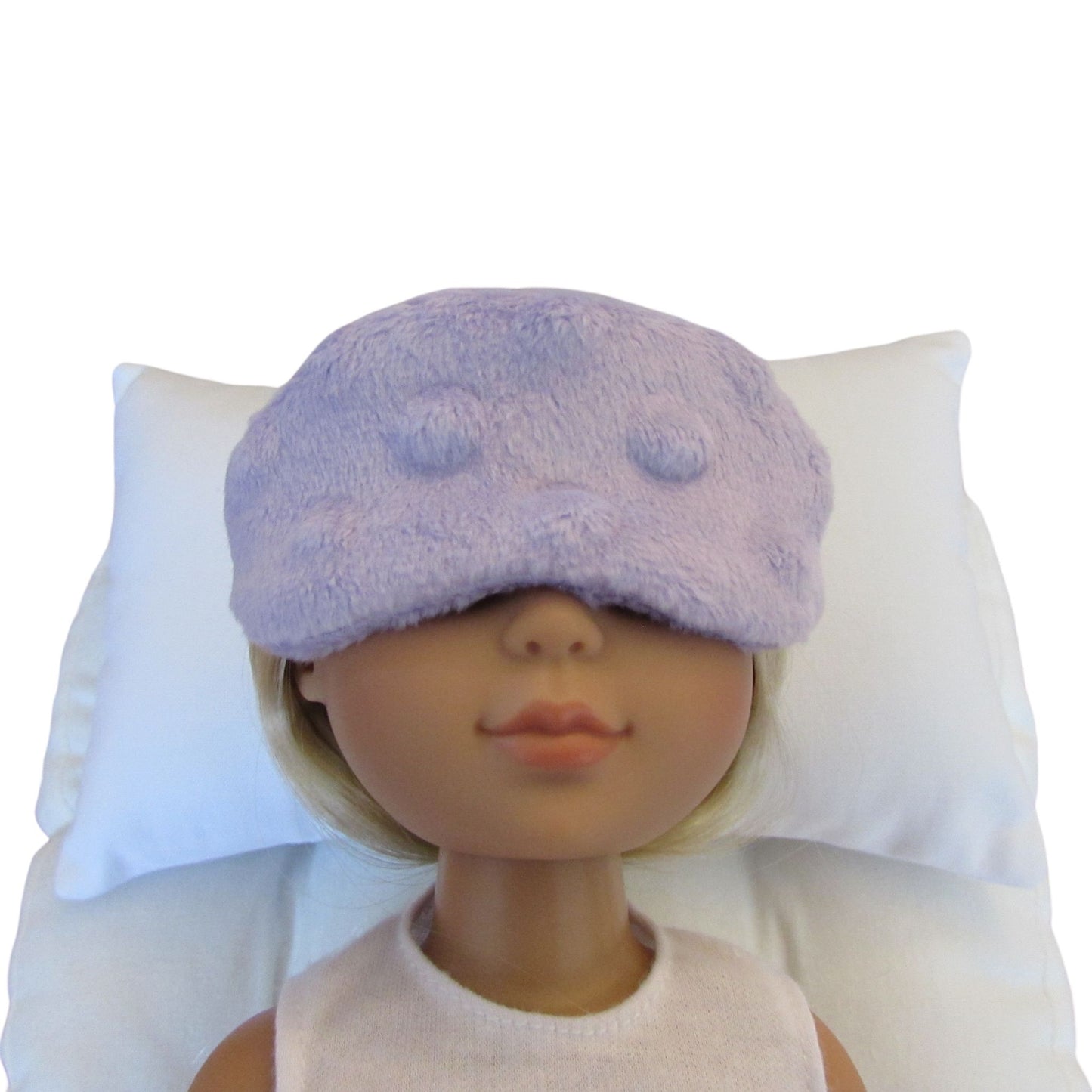 A doll with blonde hair wearing a lavender minky sleep mask, resting on a white pillow against a white background.