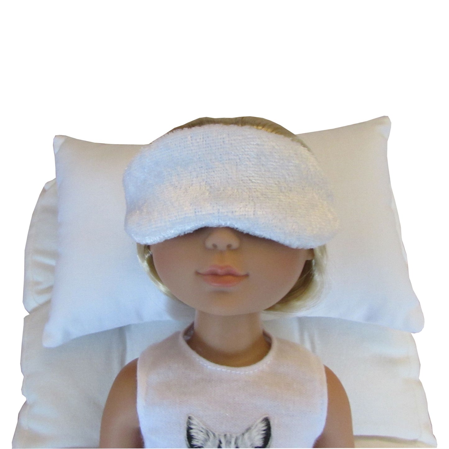 A doll wearing a white panne velvet sleep mask over closed eyes, resting on a white pillow against a white background.