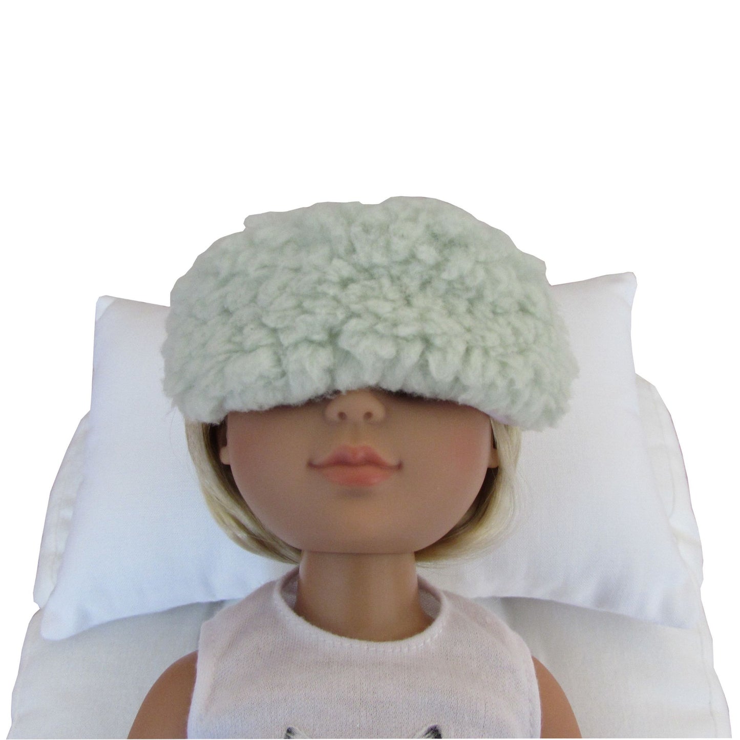 A doll with blonde hair wearing a fluffy pale mint green sleep mask, resting on a white pillow against a white background.