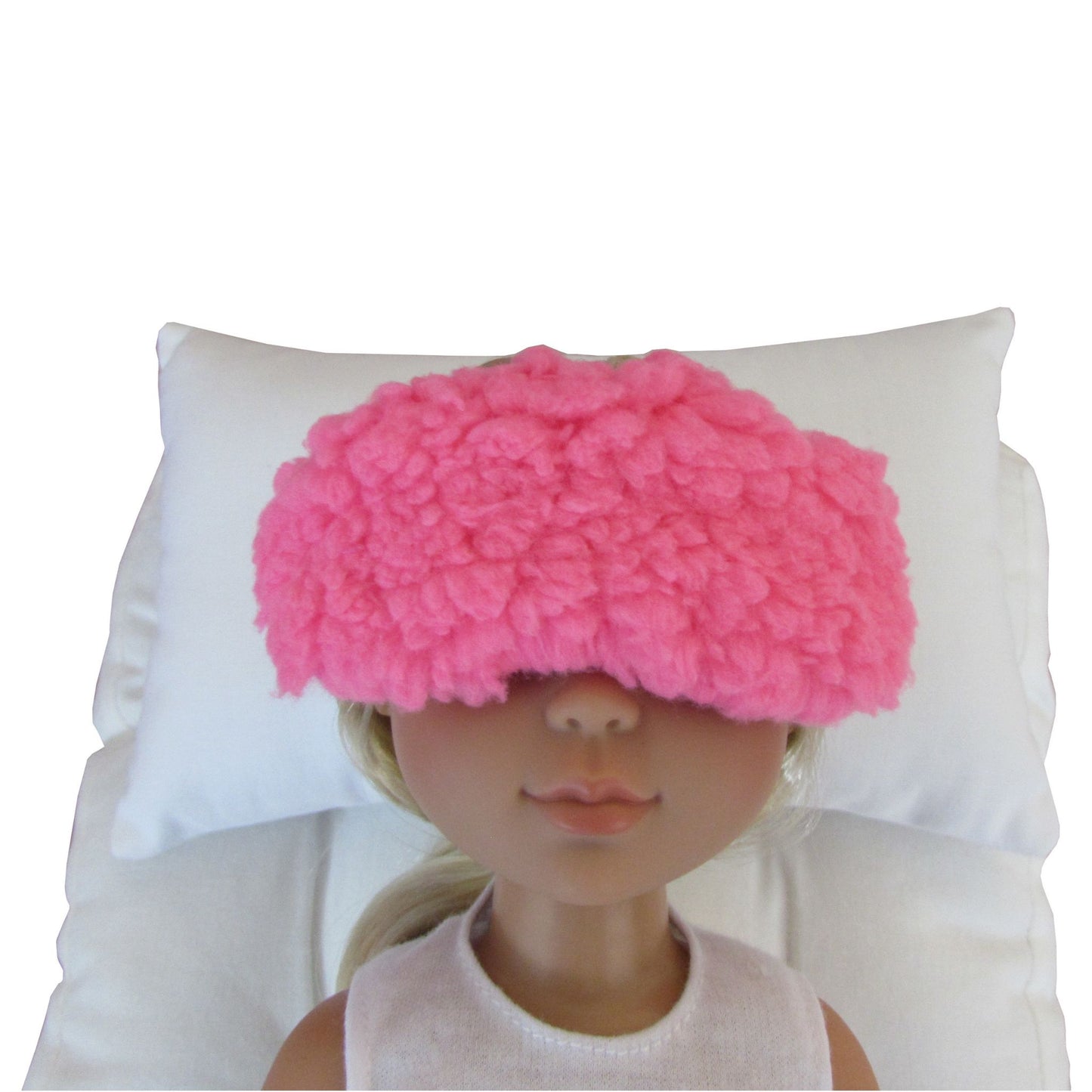 A pink fuzzy sleep mask worn by a doll, resting on a white pillow against a white background.