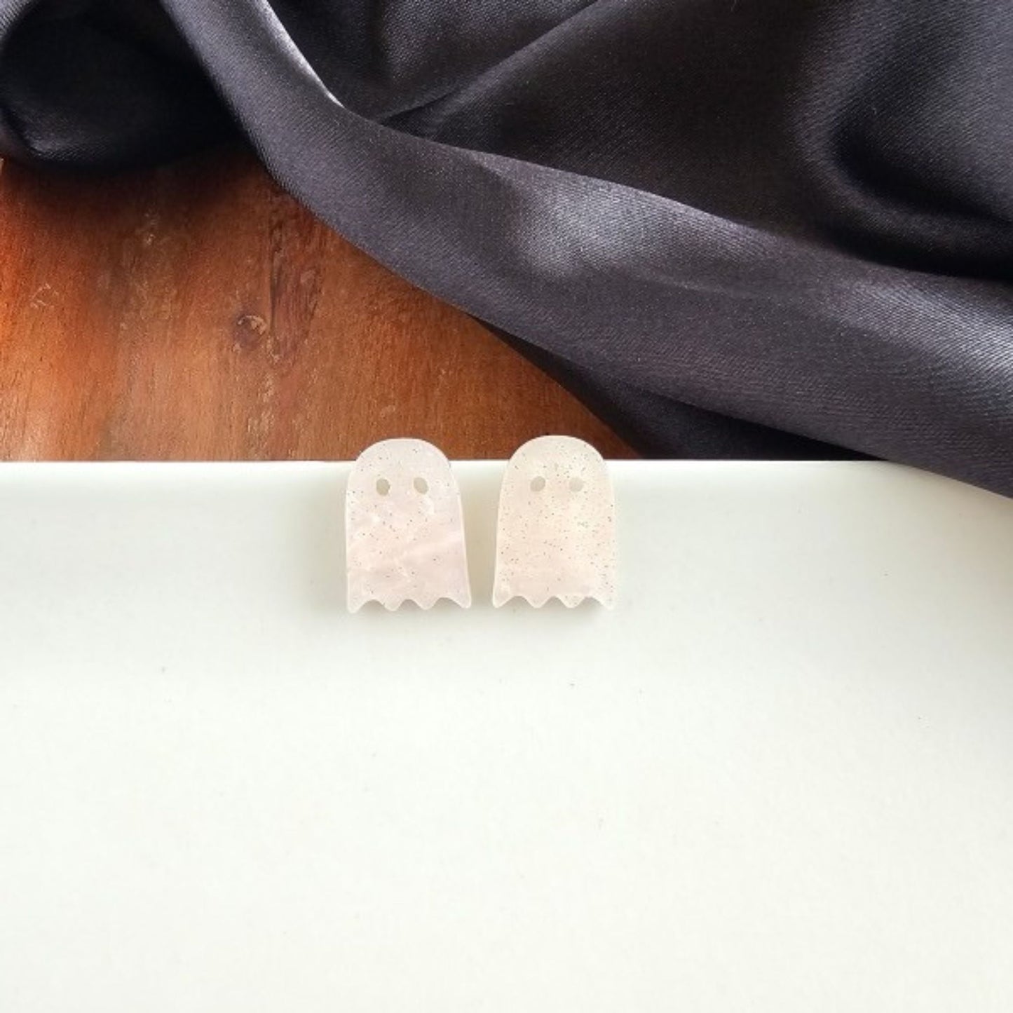 Ghost-shaped earrings on a white surface with a dark fabric background