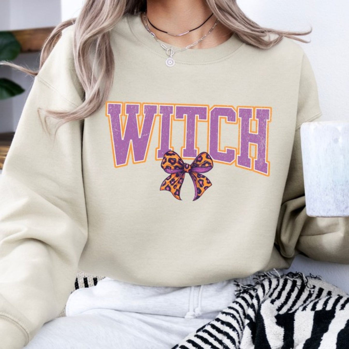 Beige sweatshirt with 'WITCH' in purple and a bow graphic, worn by a person.
