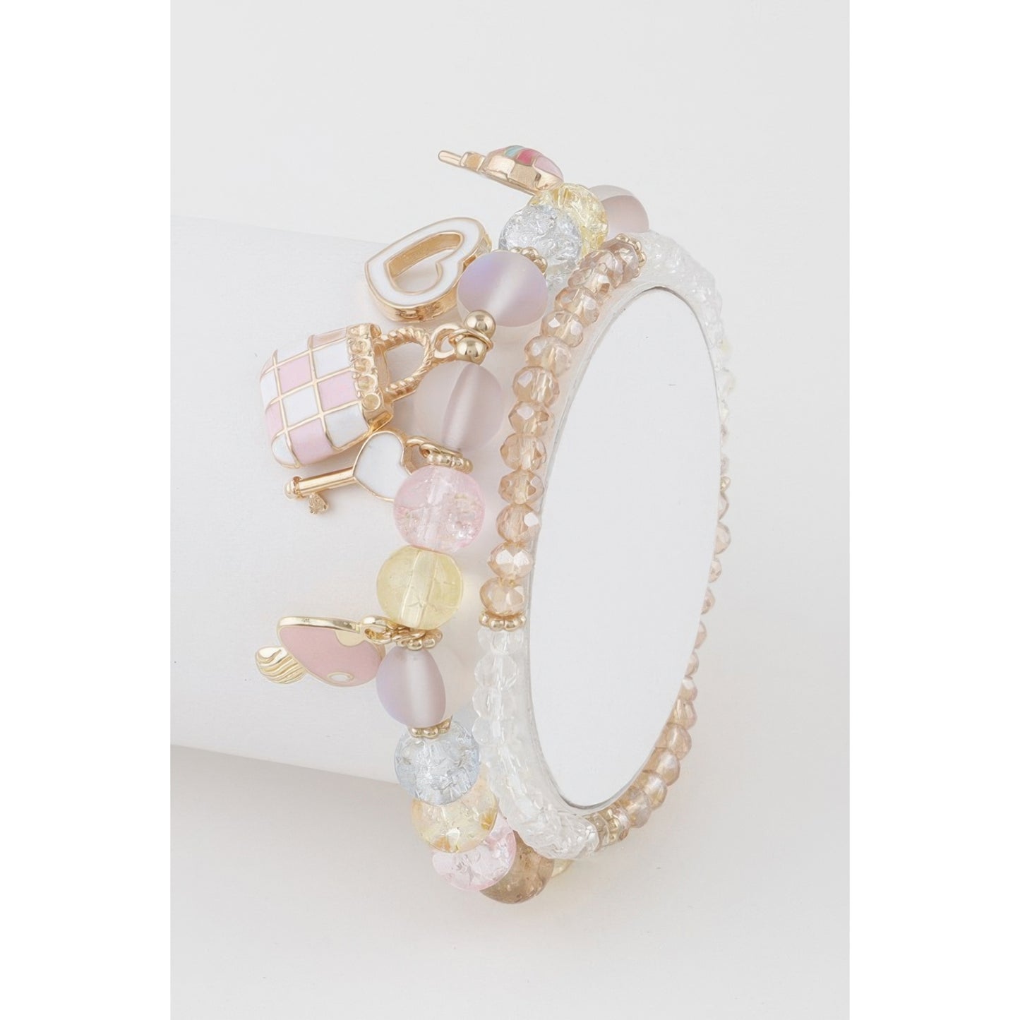 A delicate charm bracelet with pastel pink, lavender, and gold elements, displayed on a white cylindrical stand against a clean white background.