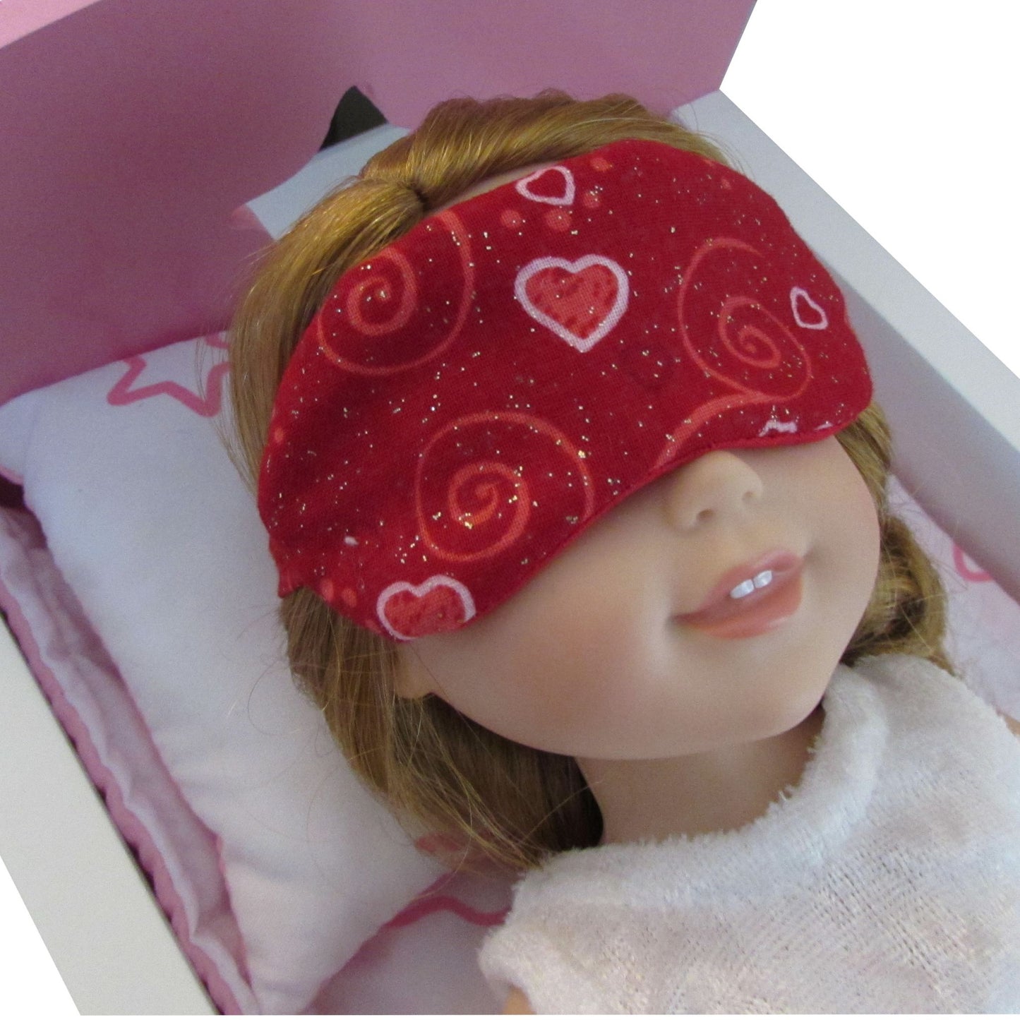 Doll wearing a red sleep mask with heart patterns on a pink bed.