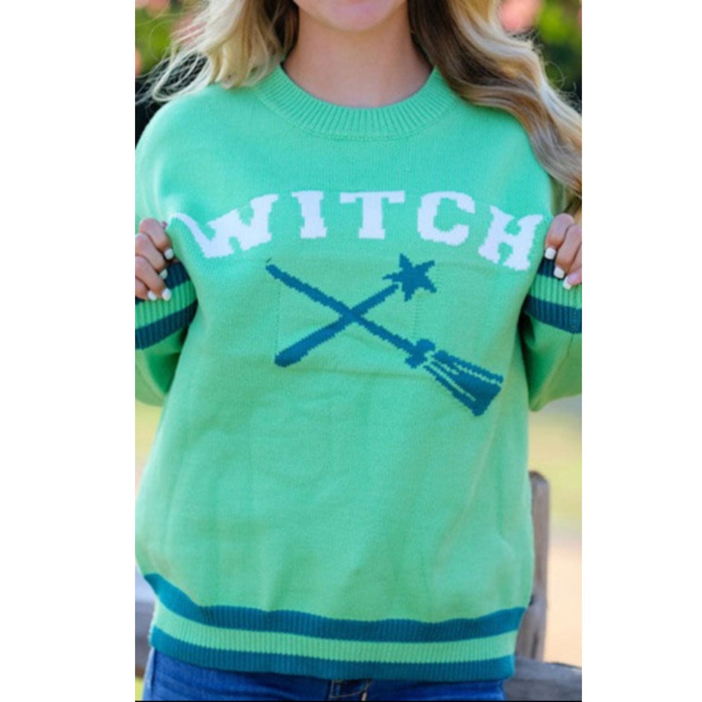 Green sweater with 'WITCH' and broomsticks printed on it, worn by a person.