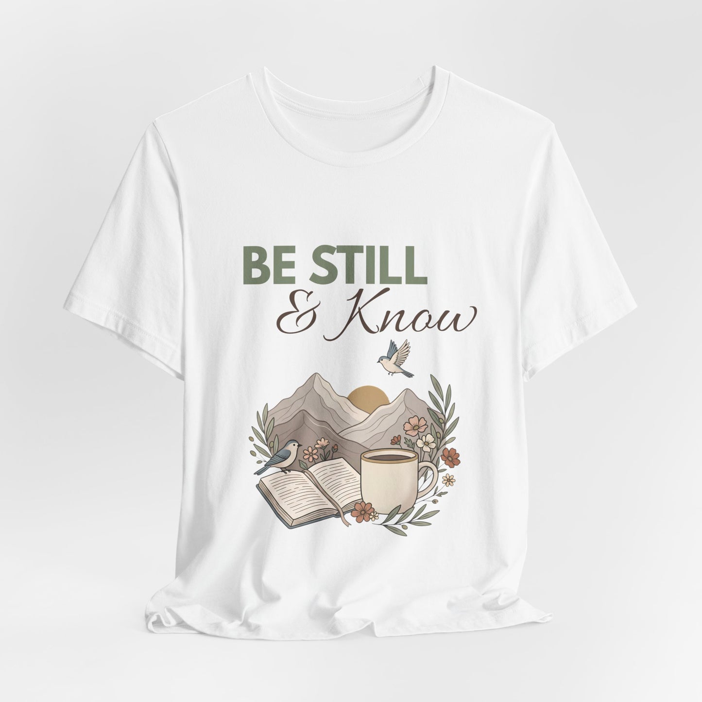 White t-shirt with 'Be Still & Know' graphic design featuring a mountain, book, and cup on a white background.