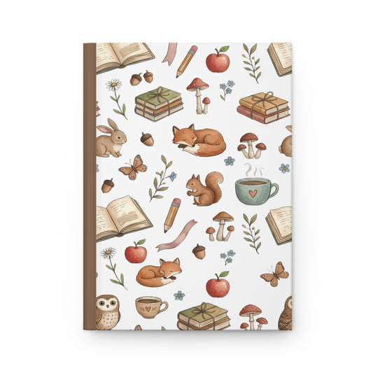 A hardcover notebook with a white cover decorated with a whimsical autumn and school-themed pattern featuring foxes, rabbits, mushrooms, acorns, books, pencils, and a coffee cup, set against a clean white background.