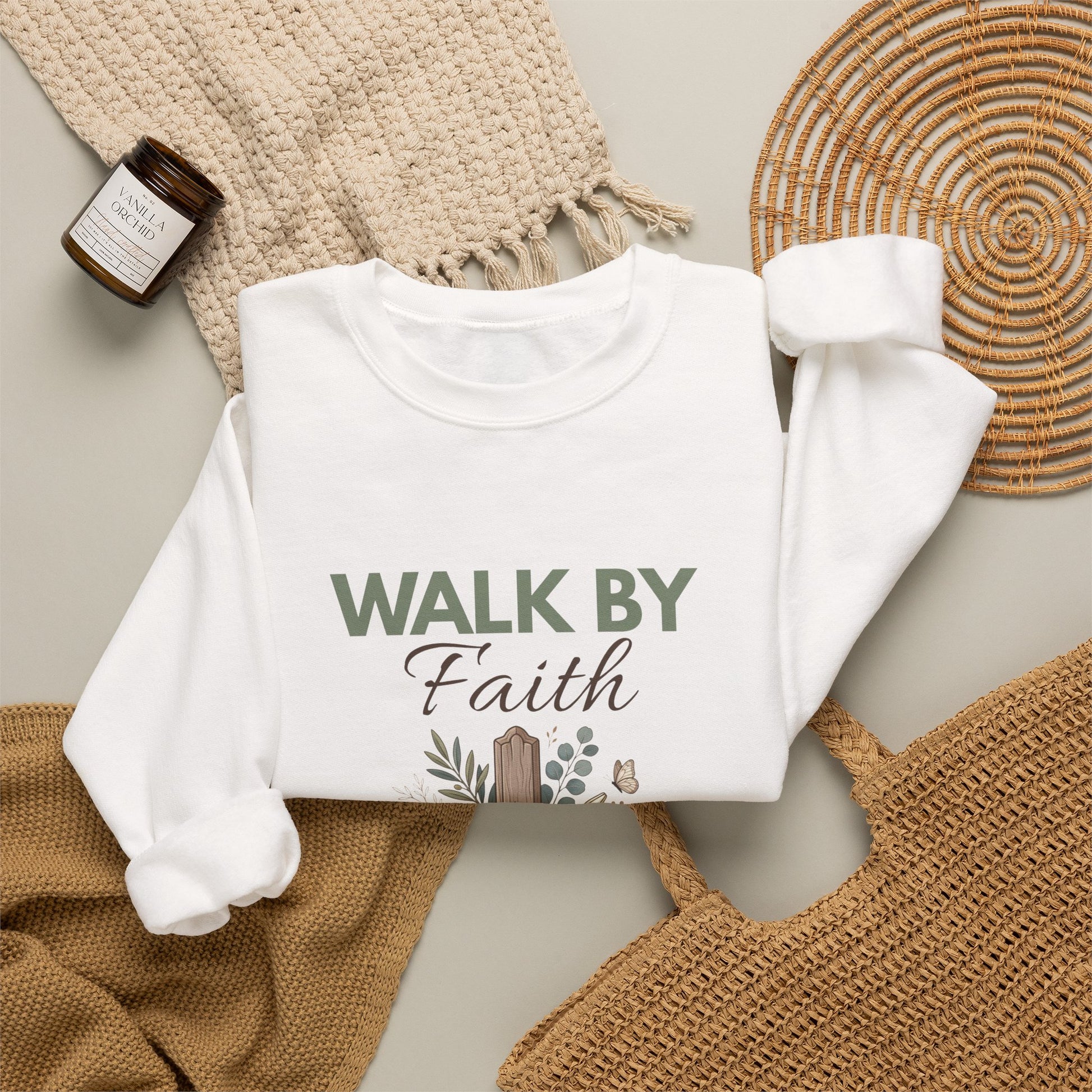 A folded white crewneck sweatshirt with 'WALK BY Faith' on the front and surrounded by a straw bag, candle, scarf, and blanket.