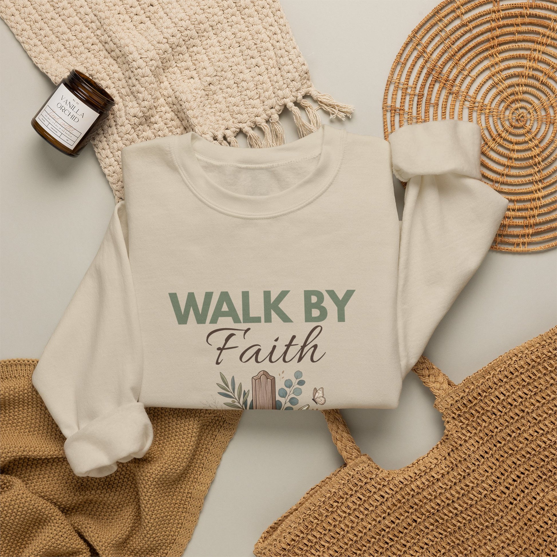 A flat lay of a sand colored crewneck sweatshirt with 'WALK BY Faith' on the front surrounded by a straw bag, a blanket,  a candle, and a scarf.