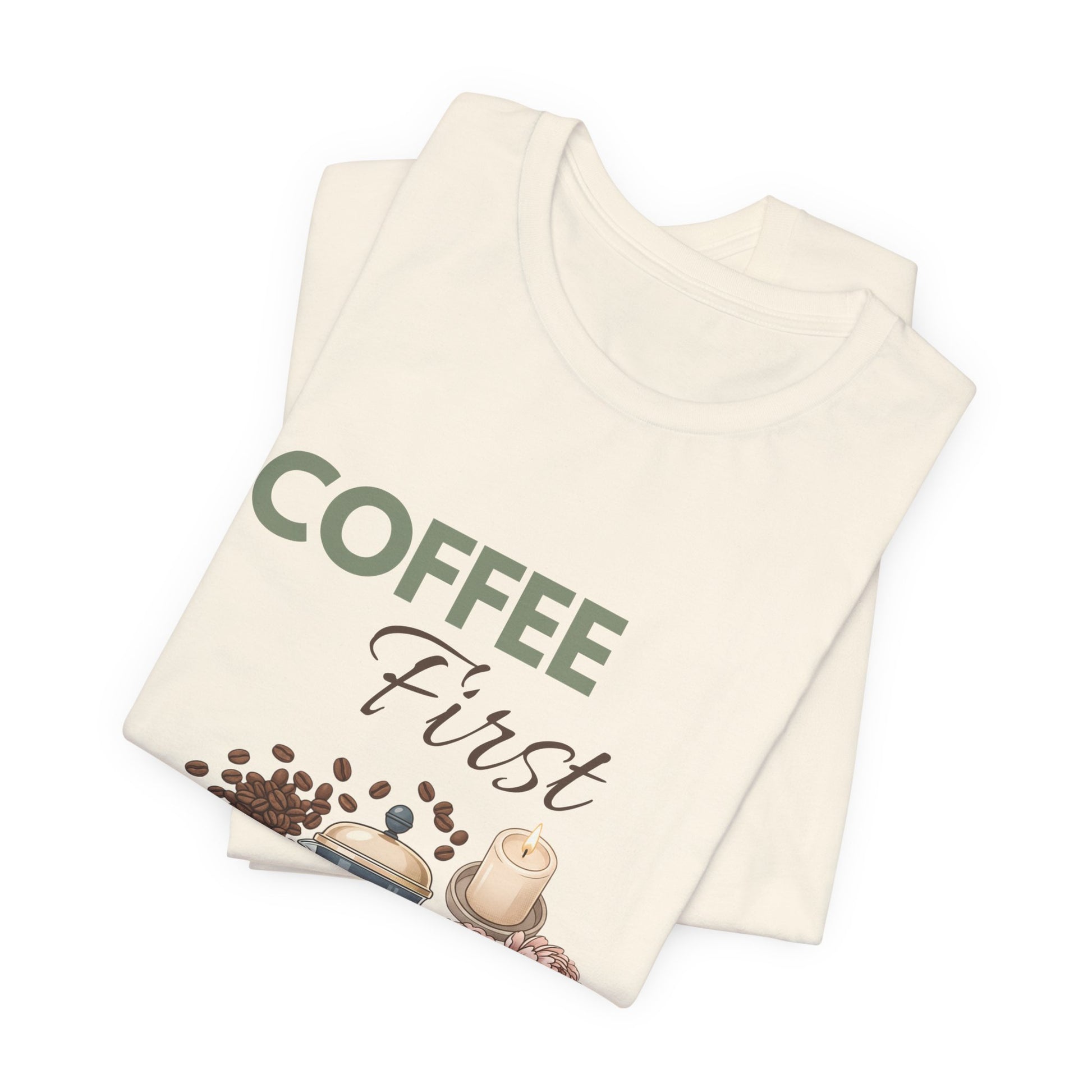 Folded natural colored t-shirt with 'Coffee First' text and coffee-themed graphics on a white background