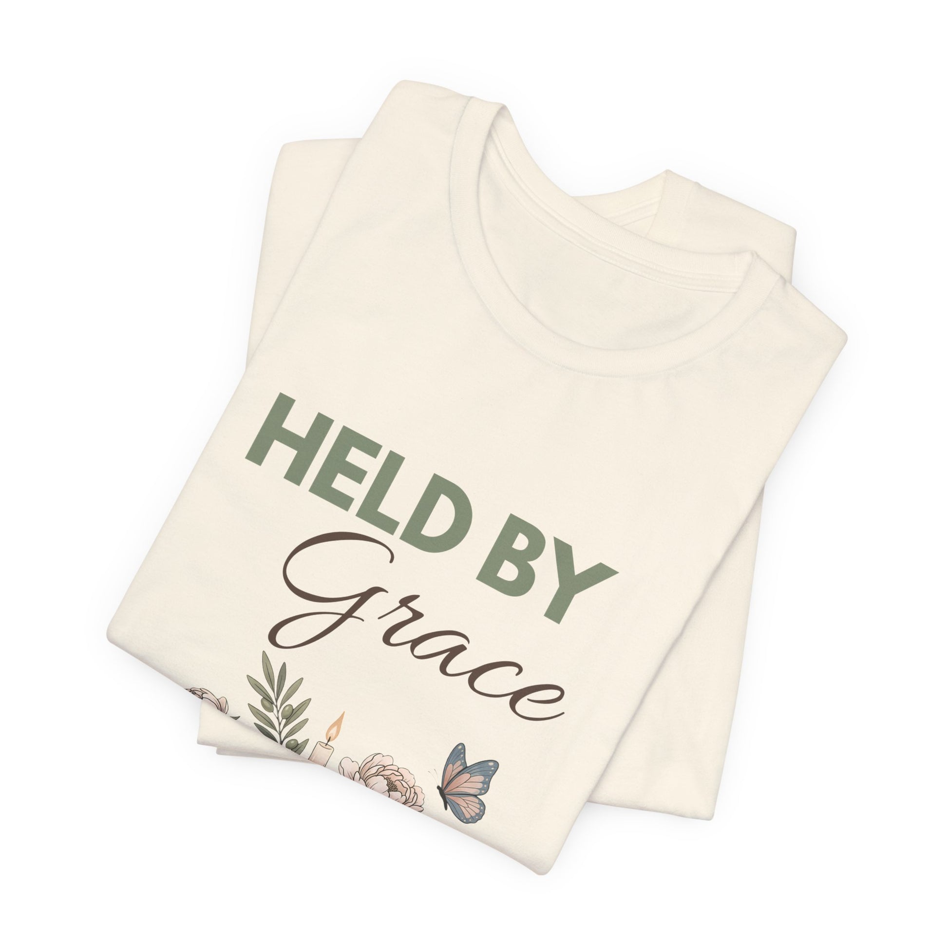 Folded natural colored t-shirt with 'HELD BY Grace' text on a white background