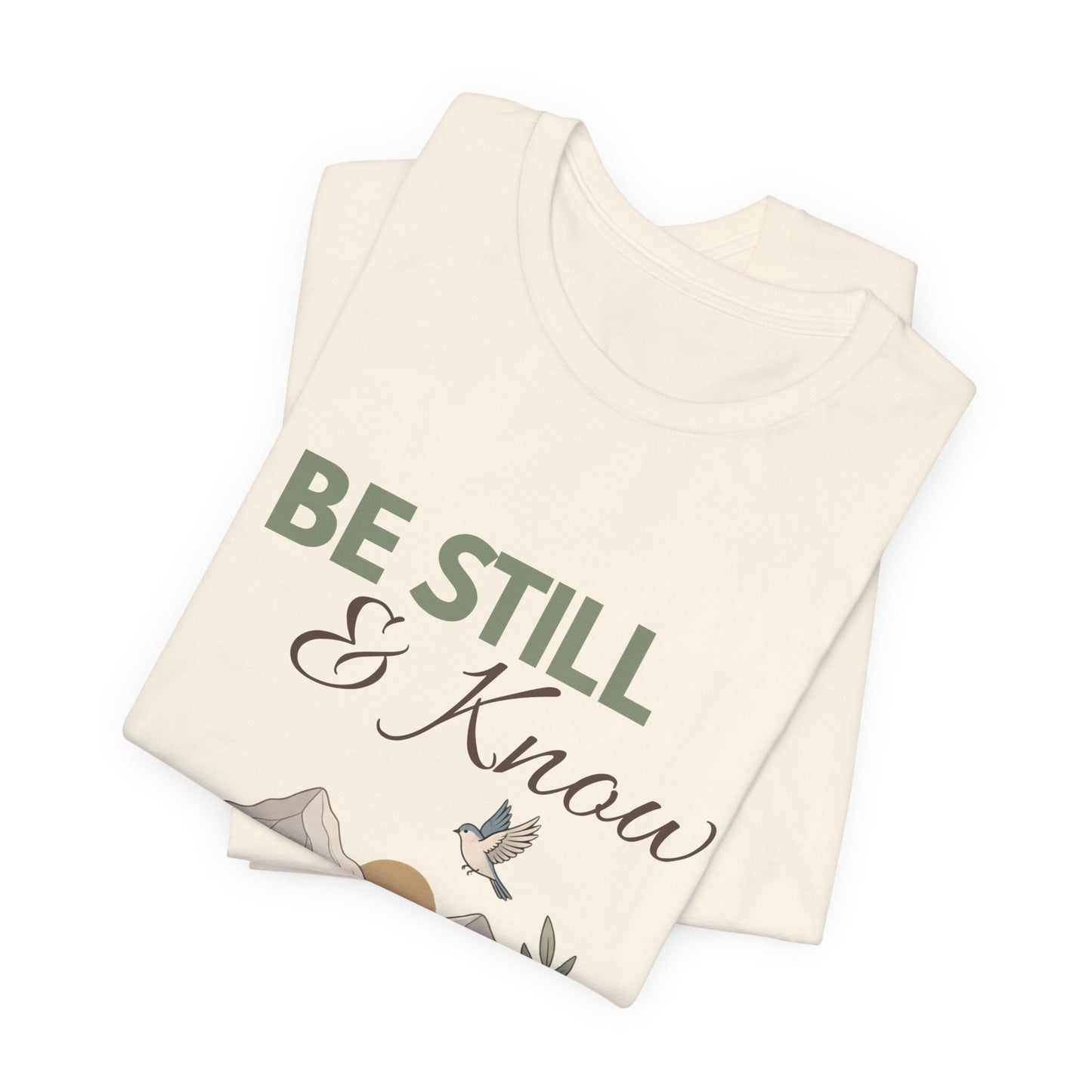 Folded natural colored t-shirt with 'Be Still & Know' text on a white background