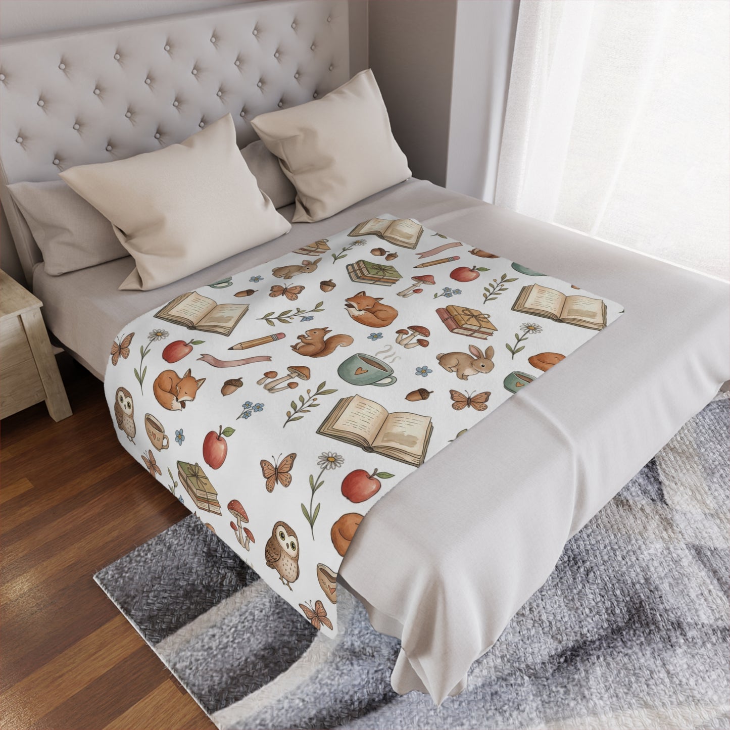 A cozy bed in a bedroom setting, styled with a white minky blanket featuring a whimsical autumn forest pattern of foxes, squirrels, owls, butterflies, acorns, and open books. The bed is dressed in neutral beige sheets and pillows, set against a tufted headboard, wooden floor, and a gray patterned rug.