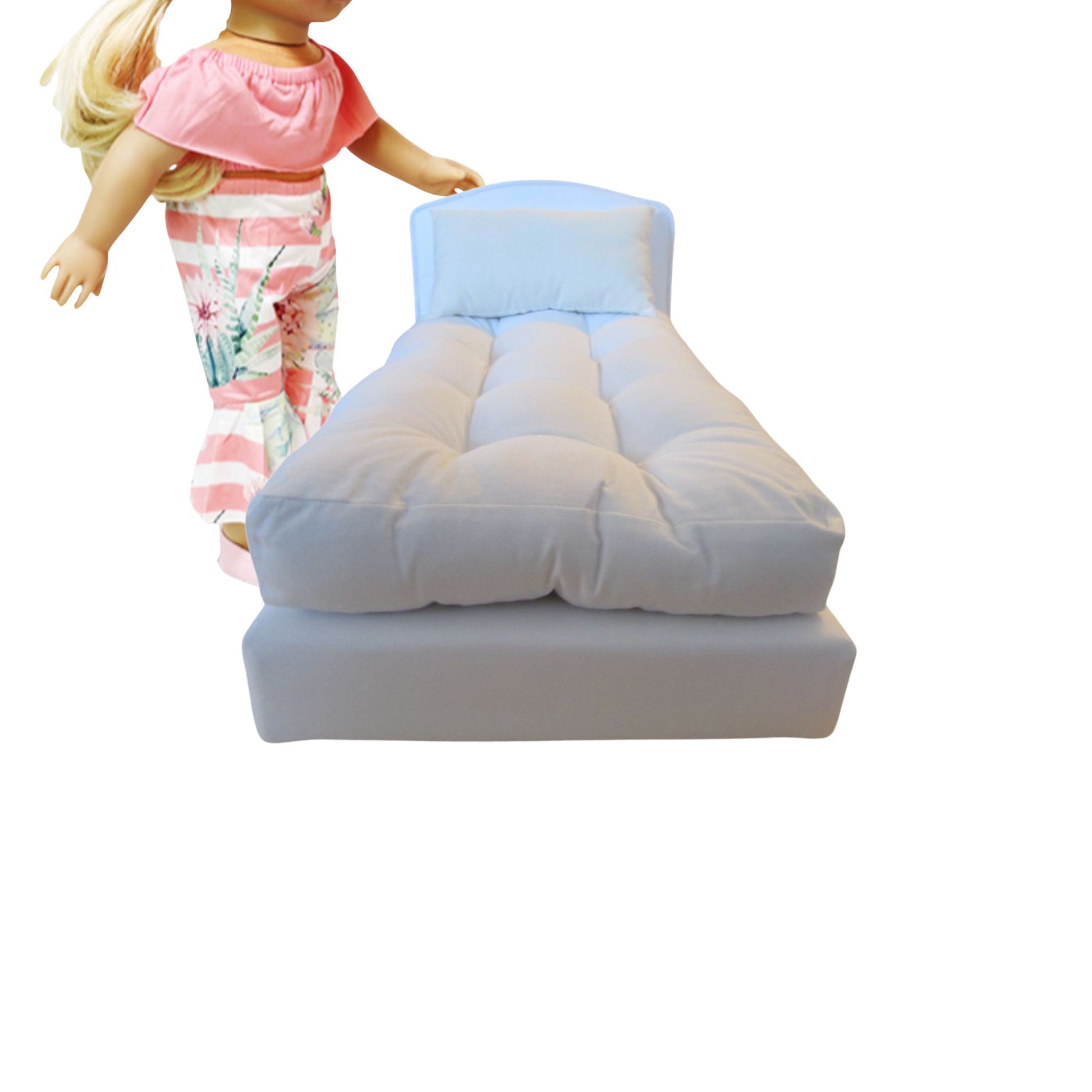 Dolll bed with white mattress next to a doll wearing a pink top and floral pants on a white background