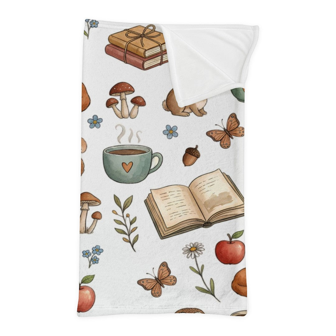 A white minky sleeping bag with a corner folded down, featuring a whimsical watercolor pattern of mushrooms, a coffee mug, an open book, butterflies, acorns, and red apples, shown on a clean white background.