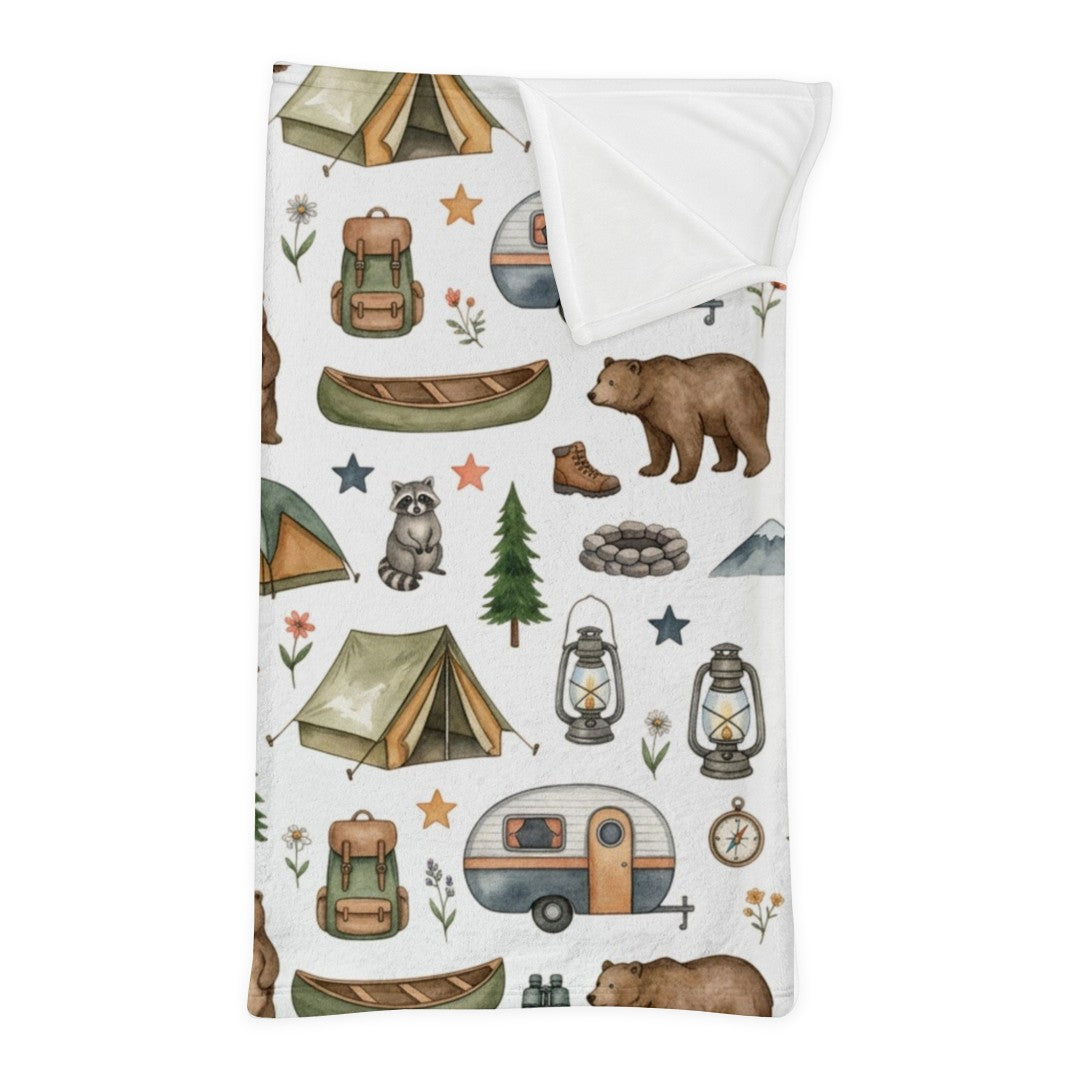 A white minky sleeping bag with a corner folded down, with a white reverse side, featuring a whimsical pattern of tents, campfires, lanterns, a canoe, bears, a raccoon, trees, stars, and cabins on a white background.