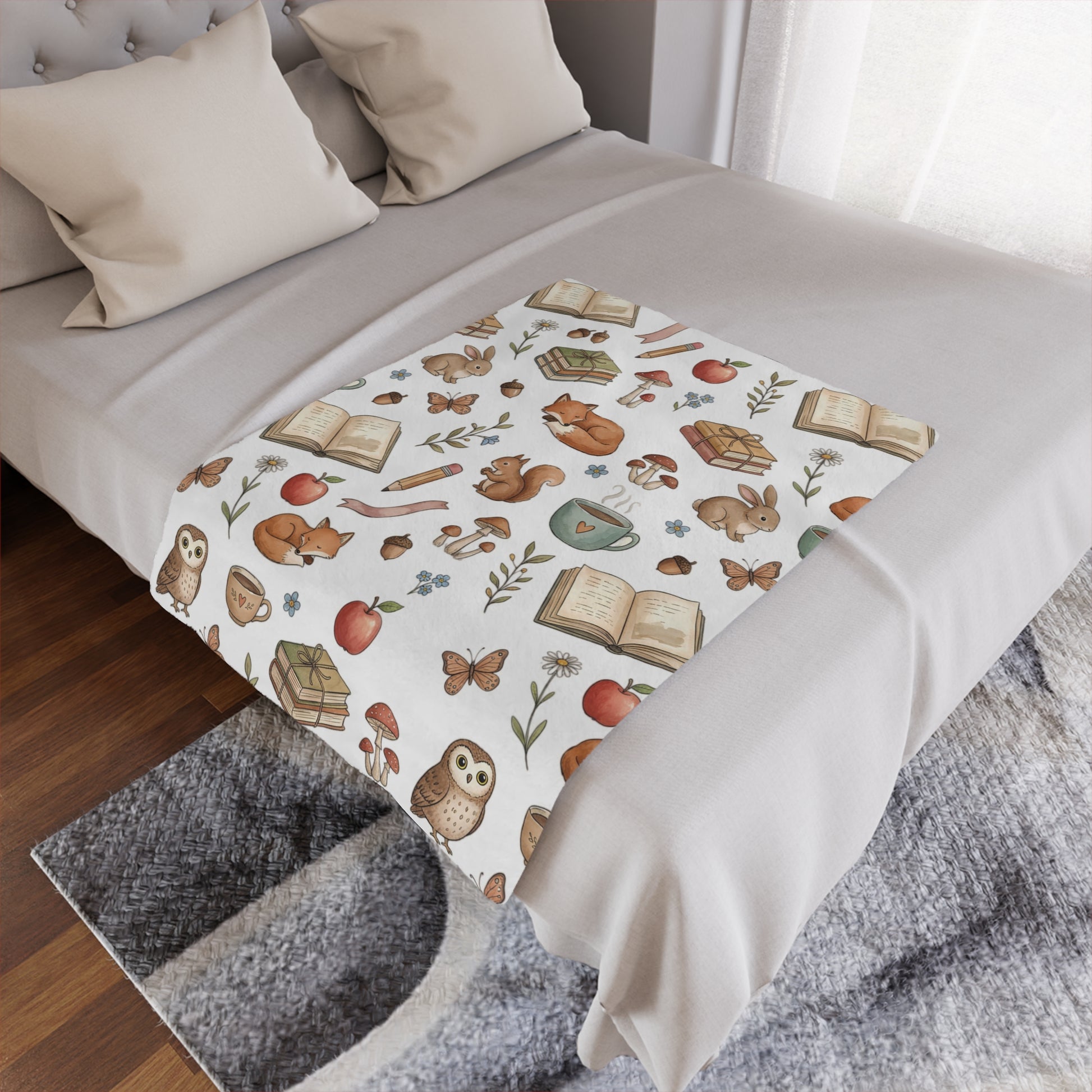 A white throw blanket with a whimsical autumn woodland pattern featuring owls, squirrels, acorns, and books, styled on a bed with neutral bedding.