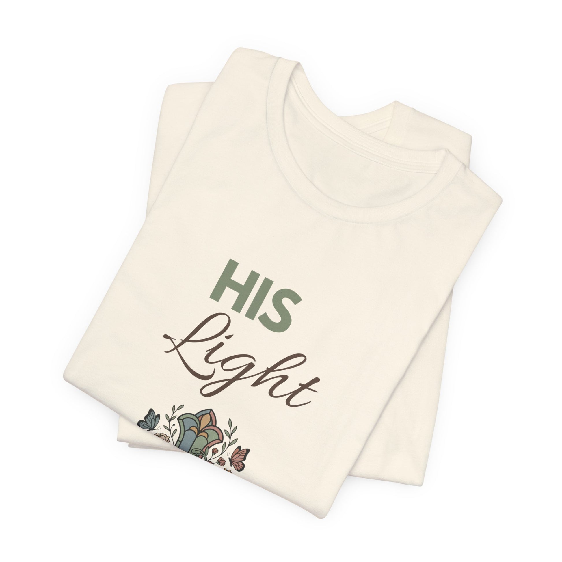 Folded natural colored t-shirt with 'His Light' text and floral design on a white background