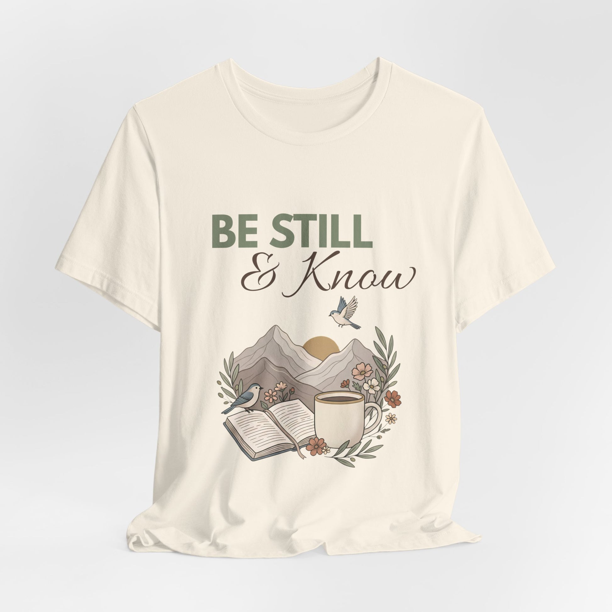 A natural colored t-shirt with 'Be Still & Know' graphic design on a light gray background
