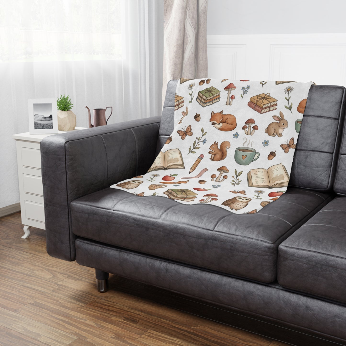 A cozy minky blanket with a whimsical woodland and book-themed pattern draped over a dark gray leather sofa in a bright living room setting.