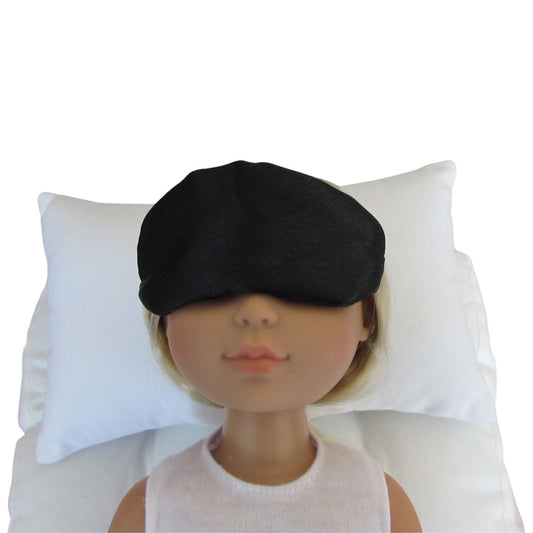 A black bridal satin sleep mask displayed on a doll's face, resting against a white pillow on a white background.