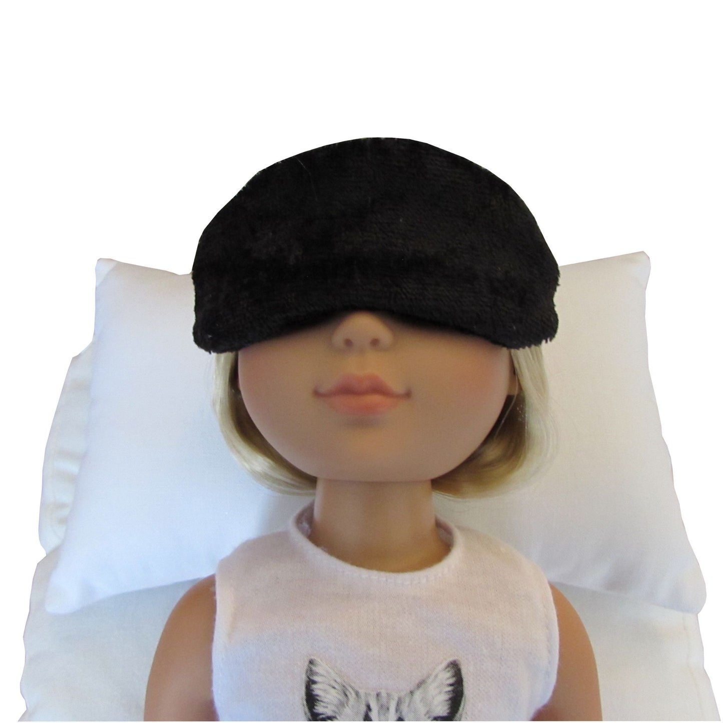 A doll with blonde hair wearing a black panne velvet sleep mask, lying on a white pillow against a white background.