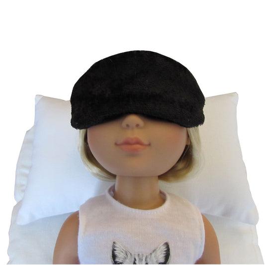 A doll with blonde hair wearing a black panne velvet sleep mask, lying on a white pillow against a white background.