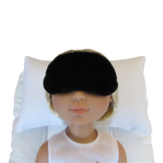 A black velvet sleep mask shown on a doll's head resting against a white pillow, demonstrating its use.
