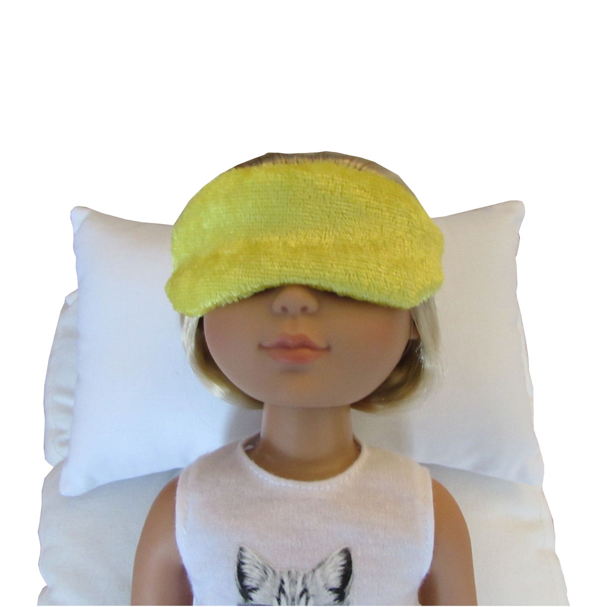 A doll with blonde hair wearing a bright yellow panne velvet sleep mask and a white sleeveless top, resting on a white pillow against a white background.