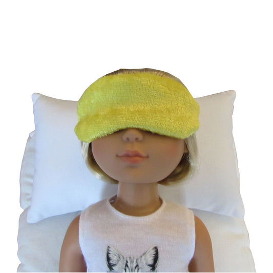 A doll with blonde hair wearing a bright yellow panne velvet sleep mask and a white sleeveless top, resting on a white pillow against a white background.
