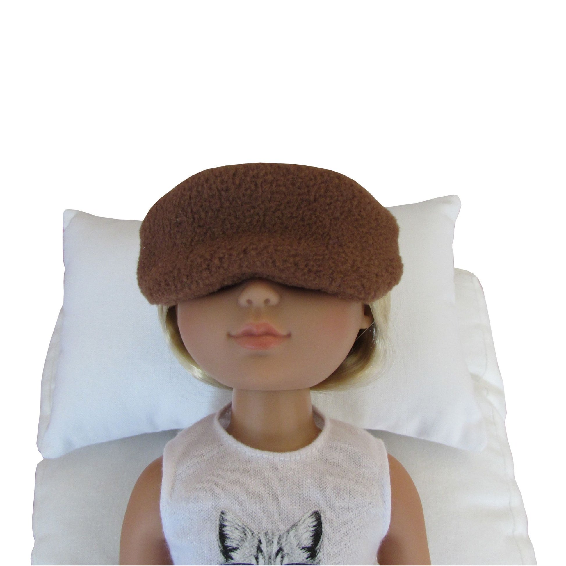 A doll with blonde hair wearing a brown fuzzy sleep mask and a white sleeveless top, lying on a white pillow against a white background.
