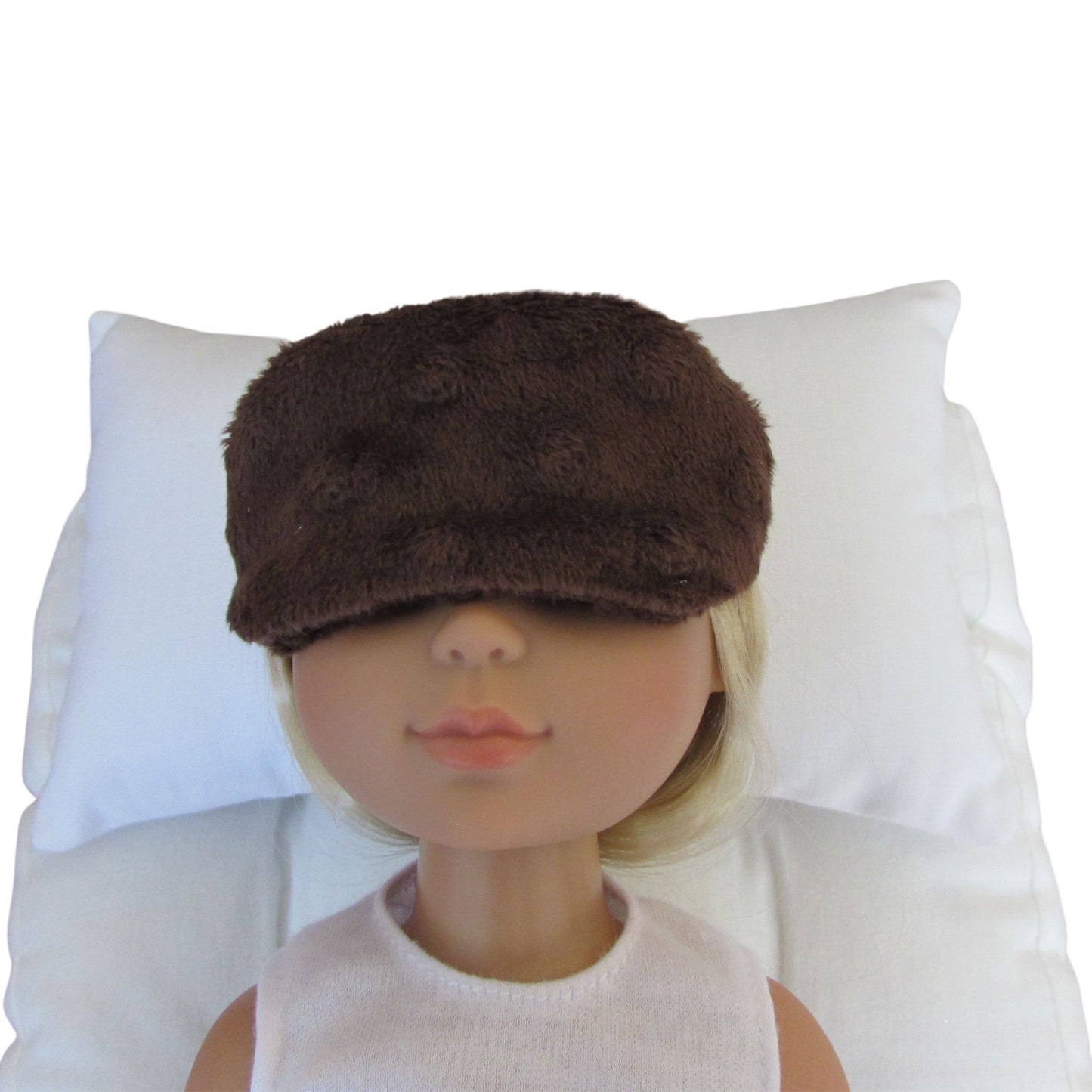 A doll with blonde hair wearing a brown minky sleep mask, resting on a white pillow against a white background.
