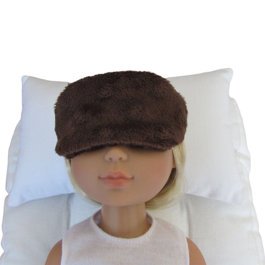 A doll with blonde hair wearing a brown minky sleep mask, resting on a white pillow against a white background.
