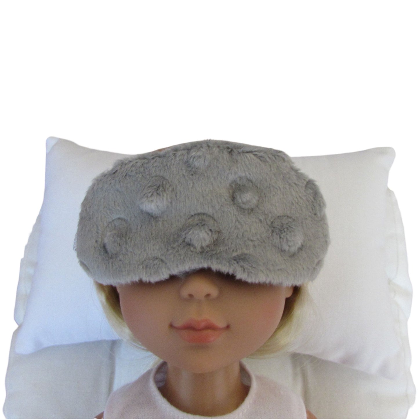 A gray minky sleep mask with raised pom-pom details, shown on a doll's face resting against a white pillow.