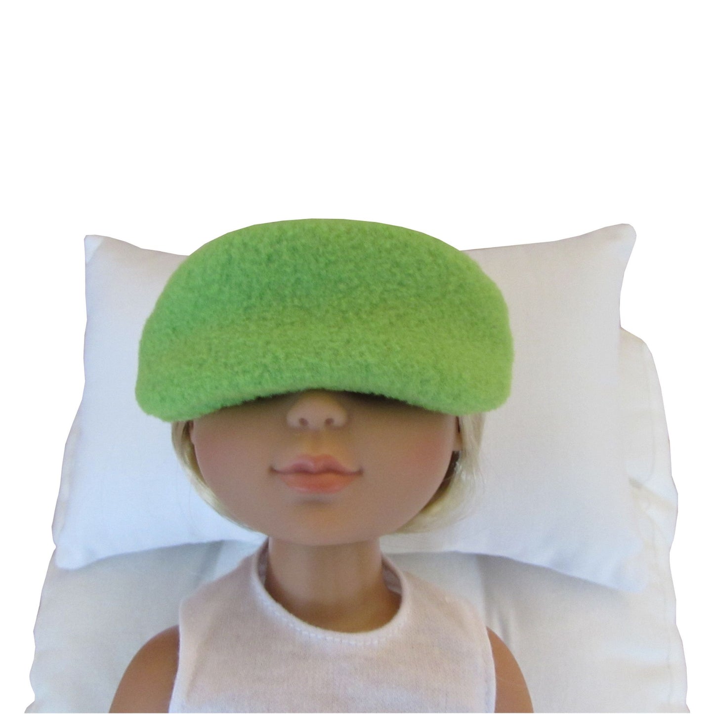 A doll with blonde hair wearing a bright green fuzzy sleep mask, lying on a white pillow against a white background.
