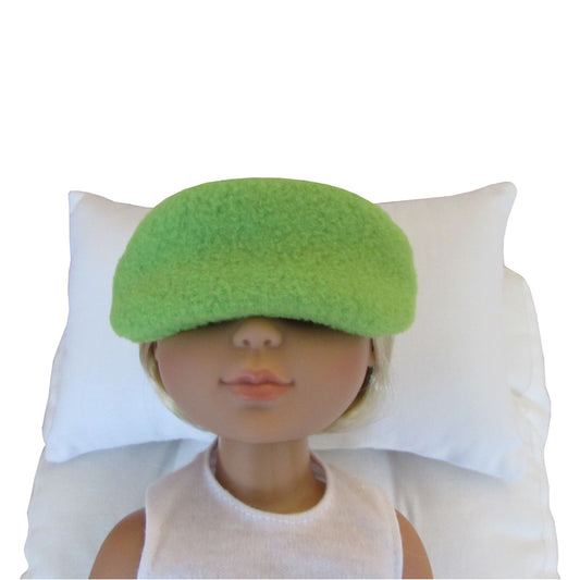 A doll with blonde hair wearing a bright green fuzzy sleep mask, lying on a white pillow against a white background.