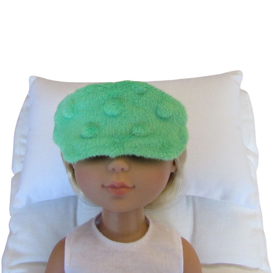 A green minky sleep mask with raised polka dots worn by a doll, resting on a white pillow against a white background.