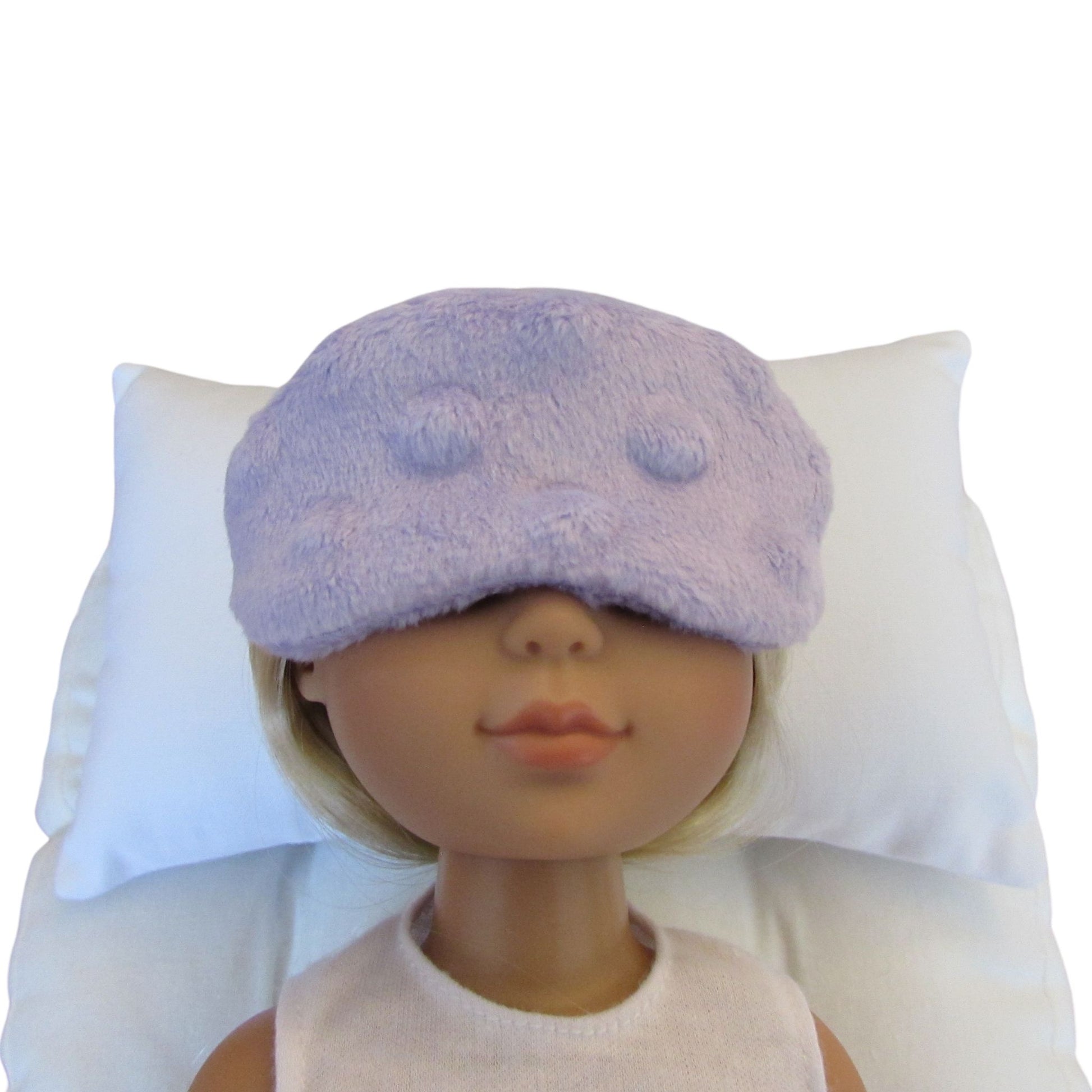 A doll with blonde hair wearing a lavender minky sleep mask, resting on a white pillow against a white background.