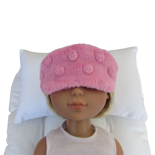 A pink minky sleep mask with raised circular dots, shown on a doll's face resting against white pillows.