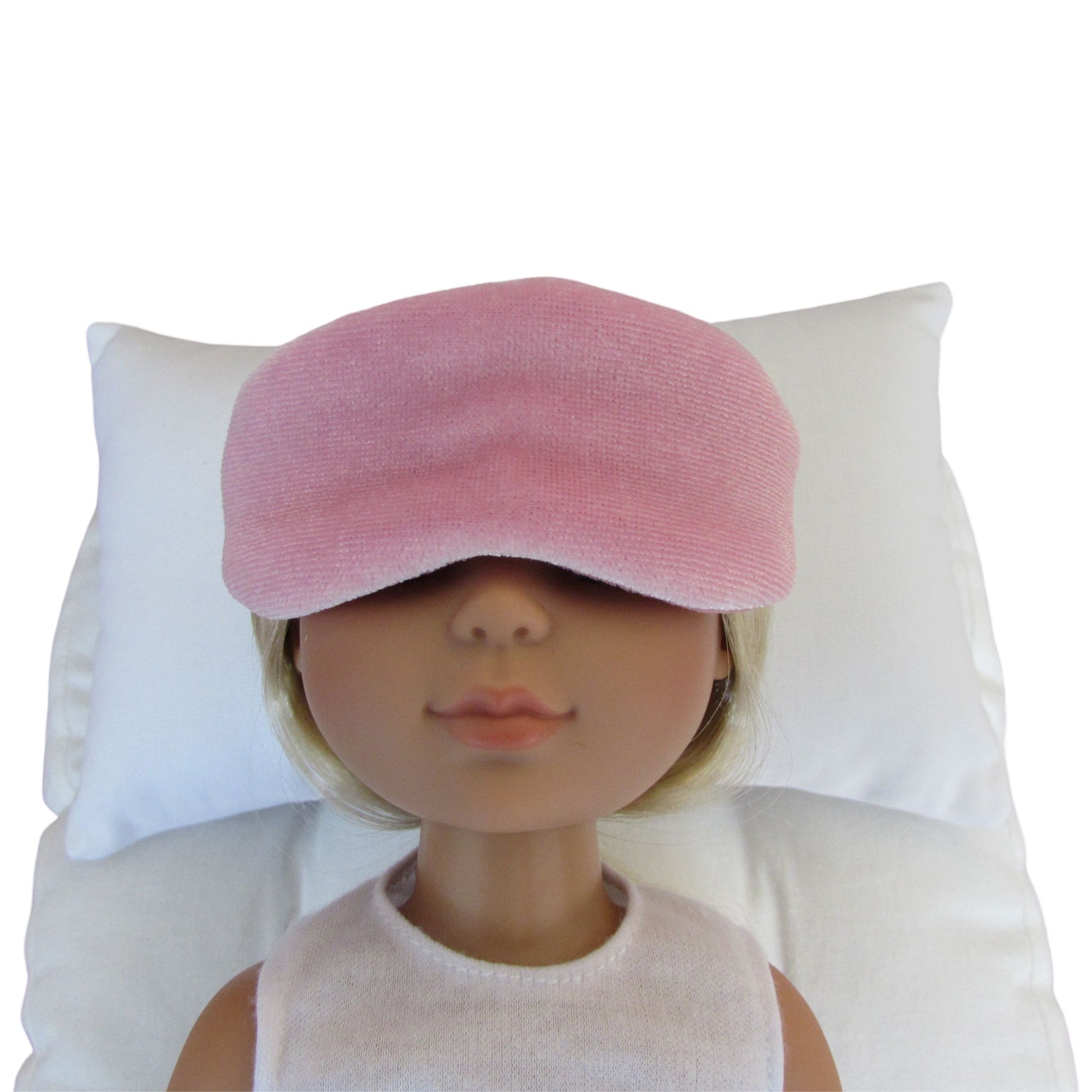 A pink stretch velvet sleep mask worn by a doll with blonde hair, resting on a white pillow against a white background.