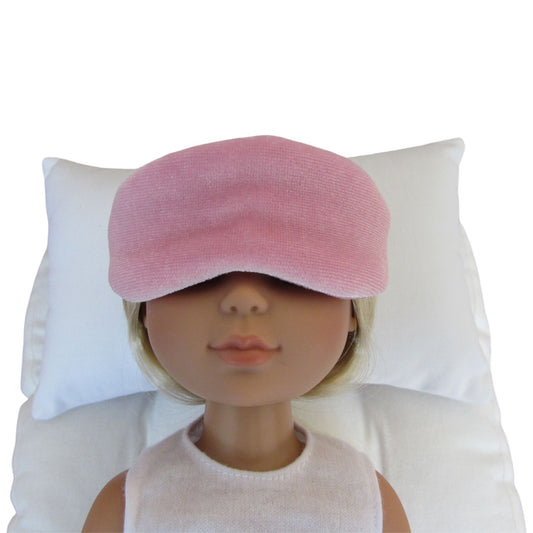 A pink stretch velvet sleep mask worn by a doll with blonde hair, resting on a white pillow against a white background.
