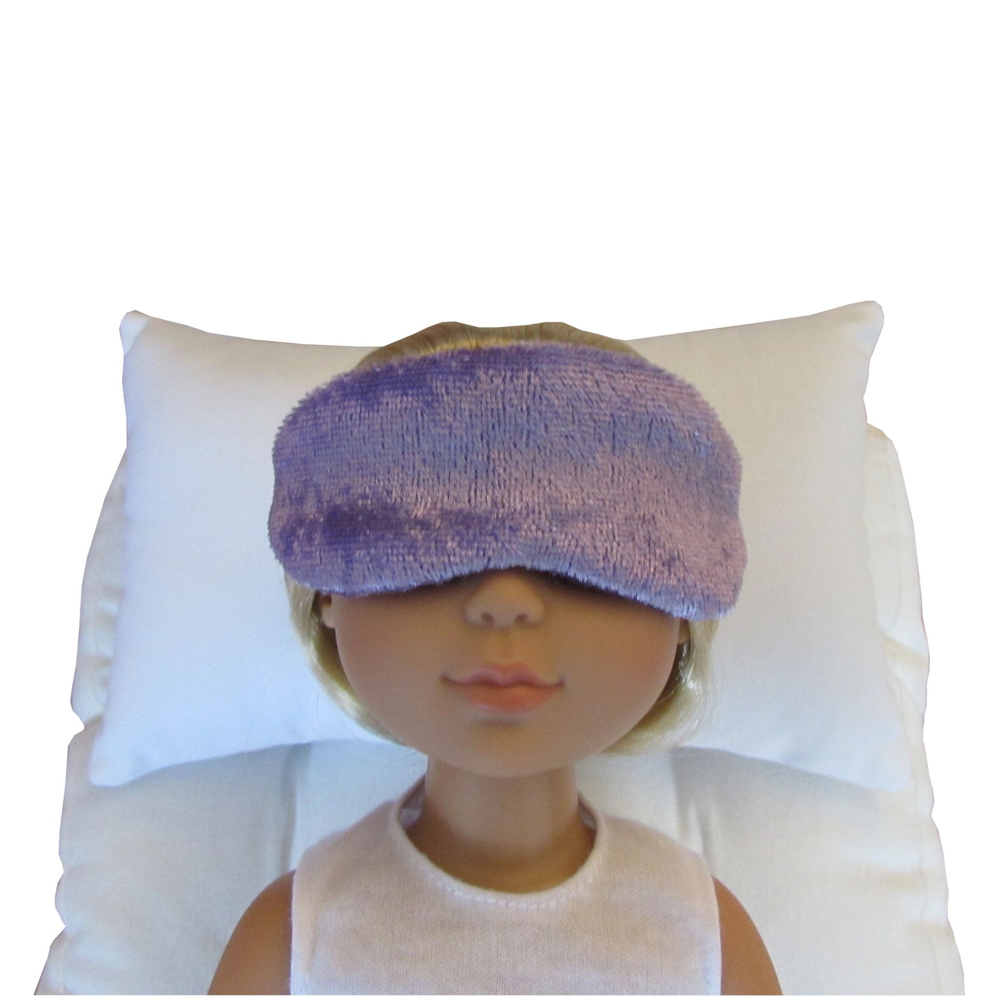 A doll with blonde hair wearing a lilac-colored sleep mask, resting on a white pillow.