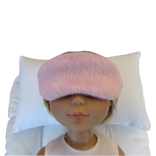 A pink panne velvet sleep mask worn by a doll, shown against a white pillow and white background.