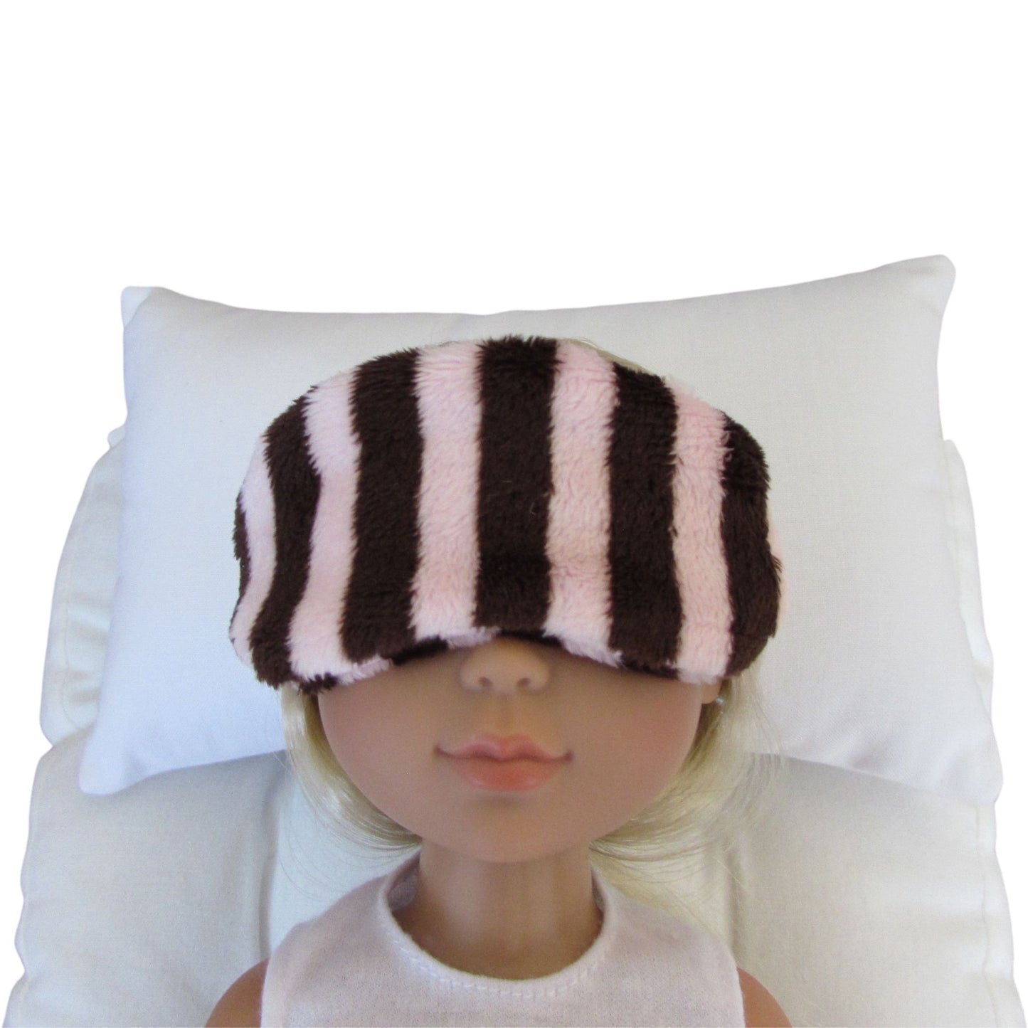A doll with blonde hair wearing a pink and brown striped minky sleep mask, resting on a white pillow against a white background.