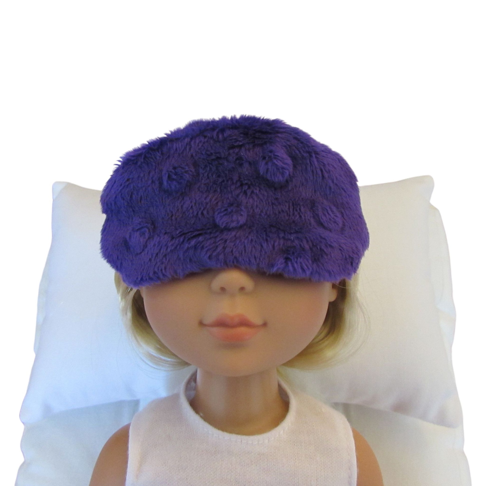 A doll with blonde hair wearing a purple minky sleep mask, resting on a white pillow against a white background.