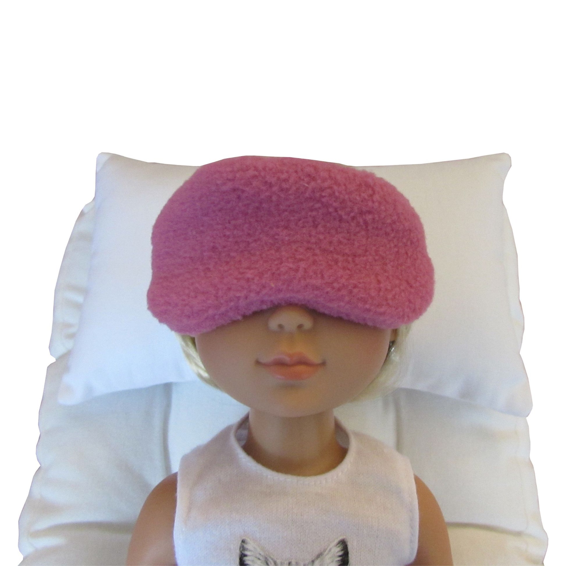 A rose-colored fuzzy sleep mask worn over a doll's eyes, resting on a white pillow against a white background.