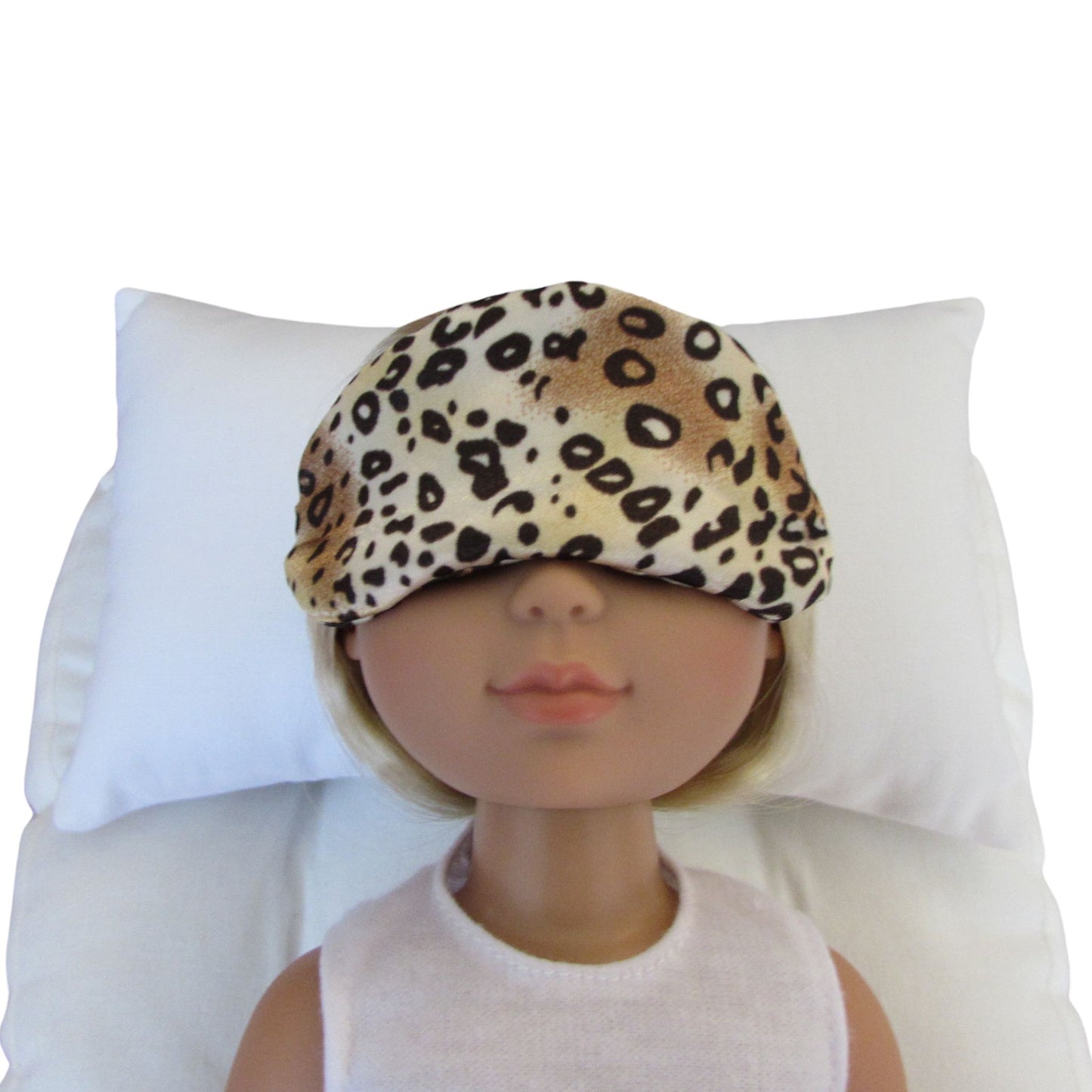 A small brown cheetah charmeuse satin sleep mask worn by a doll, resting on a white pillow against a white background.