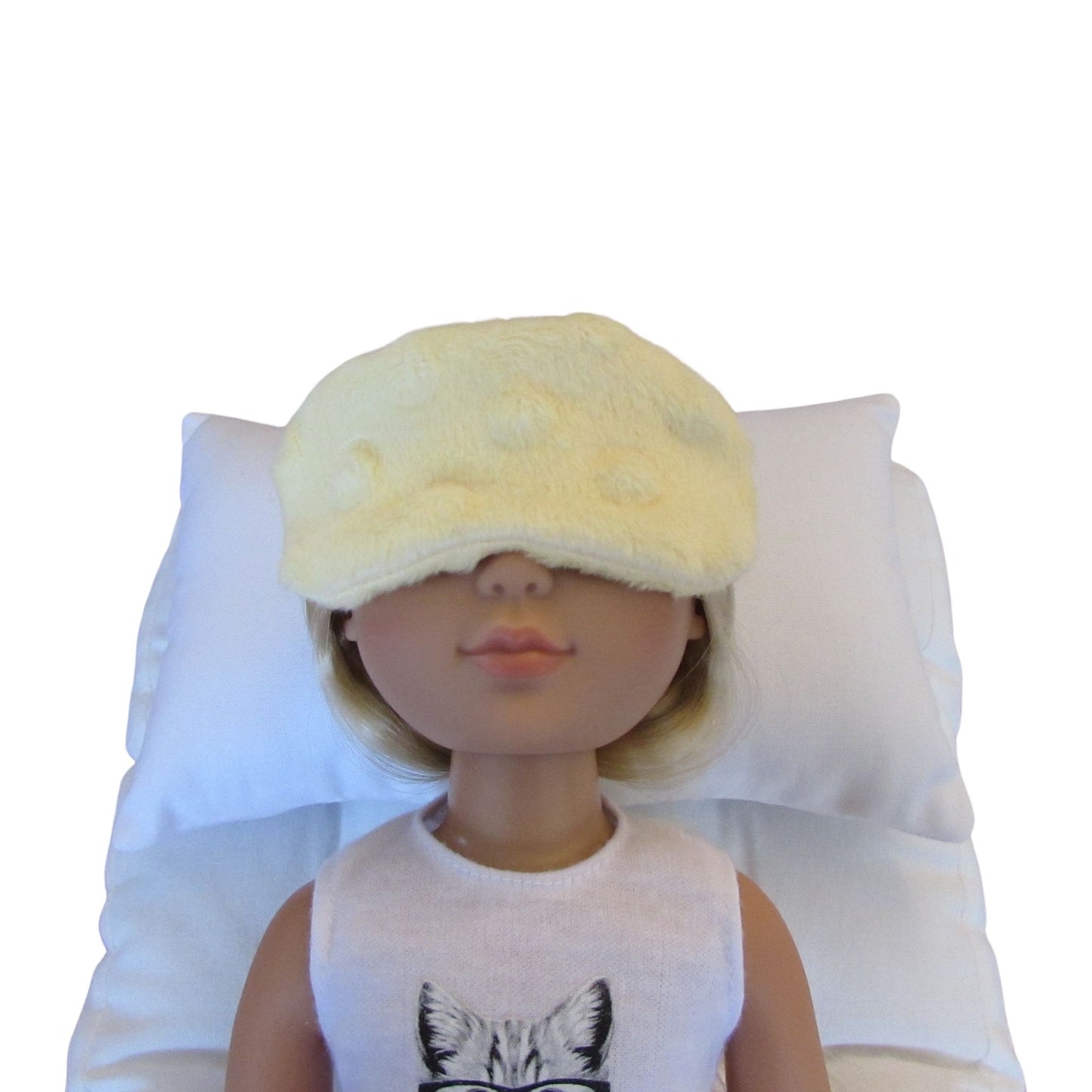 A blonde doll wearing a yellow minky sleep mask over its eyes, sitting against a white pillow on a white background.
