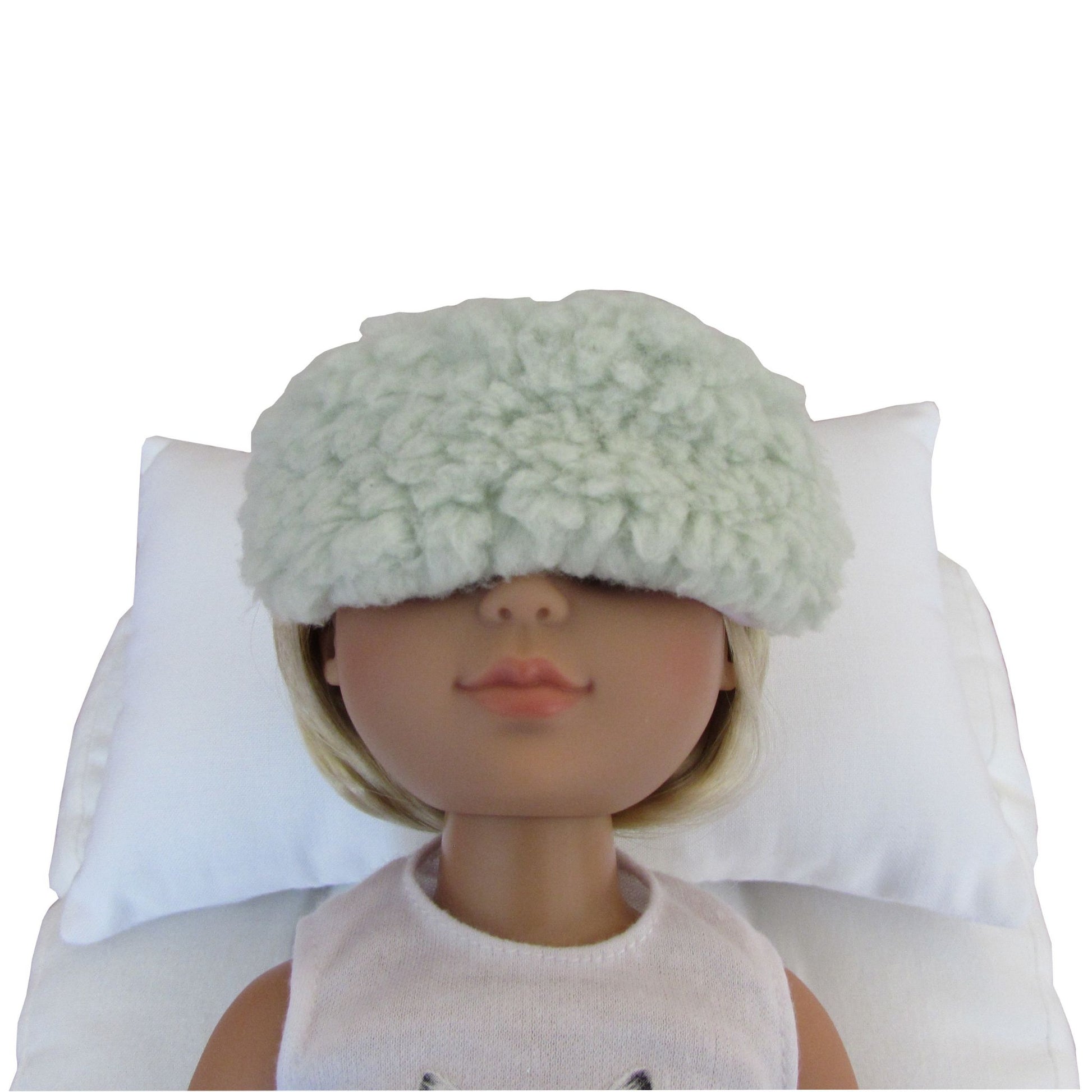 A doll with blonde hair wearing a fluffy pale mint green sleep mask, resting on a white pillow against a white background.